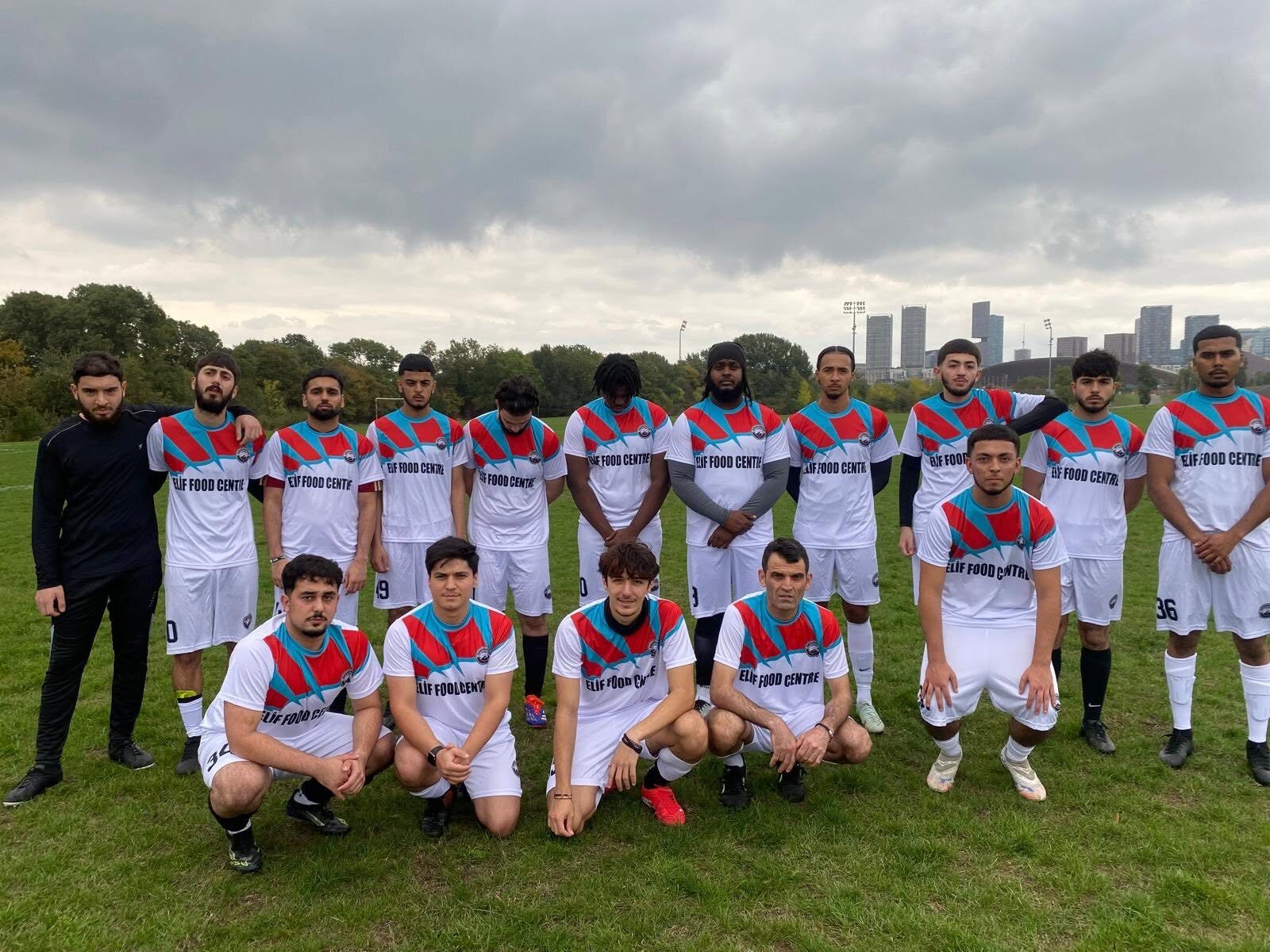 Team in white, red and blue kits lined up on a grass pitch with the London skyline behind