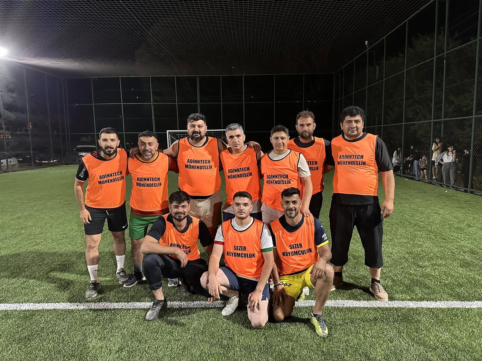 Team in orange sponsor bibs posing under the floodlights